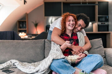 A red-haired woman is sitting on the couch, eating popcorn and watching TV, her boyfriend approaches her from behind. They smile and look happy