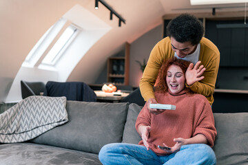Obraz premium Red-haired woman with curly hair sitting on the couch and using her phone, her boyfriend came up behind he has a present for her