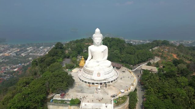 Wide, high altitude aerial view of Big Buddha in Phuket, Thailand from drone orbiting. Ocean and city in background. People visit huge Buddha templet monument for religious worship tourist attraction.