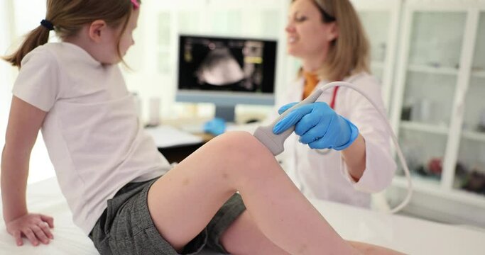 Doctor checks sore knee of little girl using ultrasound equipment. Woman in medical uniform looks at screen examining condition of joint slow motion