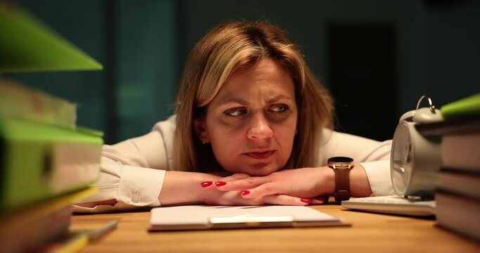 Pensive woman leans on wooden table near clipboard thinking about vacation while lightning flashing. Employee stays late at work in office feeling tired