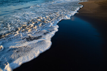 Black sandy beach with turquoise water, surf, bubbles and wave foam in warm evening light at...