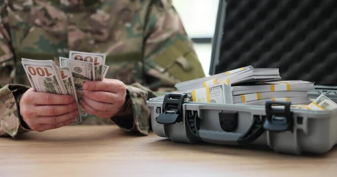 Military officer counts dollar banknotes sitting at table near open case with money stacks. Man in uniform checks cash for purchase of weapons slow motion