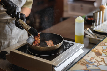 Close-up view of chef in uniform stir frying bacon in pan for cooking dish in restaurant kitchen
