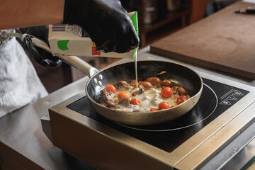 The professional chef in uniform adding cream into frying pan with dish in restaurant kitchen