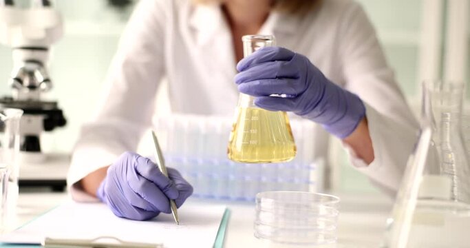 Professional scientist examines liquid oil in flask writing analysis in notebook. Laboratory assistant holds investigation of liquid slow motion