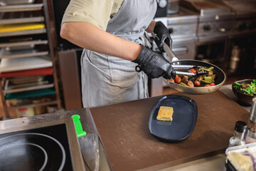 Chef putting ready dish from frying pan into the plate before serving to client in restaurant