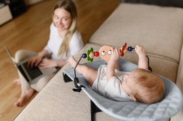 A young mother sits on the floor at home and works remotely on a laptop, a 2.5-month-old baby lies in a rocking chair and plays with a toy, focus on the child. Working mom, remote work and education