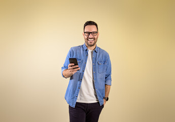 Portrait of cheerful handsome entrepreneur with hand in pocket messaging online over smart phone. Happy young man dressed in blue jeans shirt posing confidently over beige background