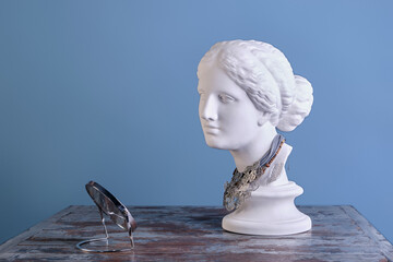 A beautiful plaster Aphrodite with different necklaces around her neck looks in the mirror, on a vintage table, on a blue background. The concept of beauty and style, self-care, time to dress up - obrazy, fototapety, plakaty