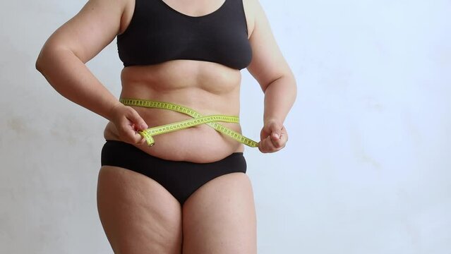 Unrecognizable fat woman wearing black underwear, tightening excessive stomach with yellow measuring tape, loosening tape on white background. Weight control, plastic surgery, liposuction, adiposity. 