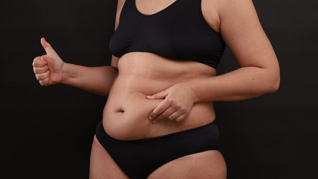 Unrecognizable buxom woman wearing black sportswear bra panties, standing on black background, caressing massaging skin of belly, showing thumb up sign, gesturing. Overweight, body positive, approval.