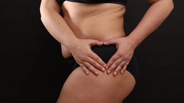 Side view of unrecognizable plus-size woman wearing black sportswear bra, standing on black background, making heart sign with hands fingers on hip panties. Health care, body positive, self-love.