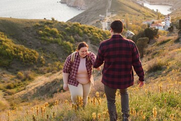 Tourists: A couple travels through the spring mountains. A man in a plaid shirt gives his hand to help a plump woman up the mountain. Walks in the open air. Travel and active life concept. Travel life