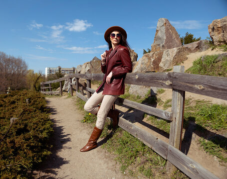 A young American woman in a hat and leather jacket stands on the road near rocks and a wooden fence. woman with glasses looking into the distance