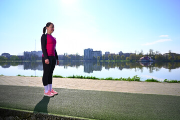 A young woman exercising at a tranquil lake during a beautiful morning. She's wearing workout clothes and appears focused and relaxed as she performs her exercise routine. Natural stunning backdrop.