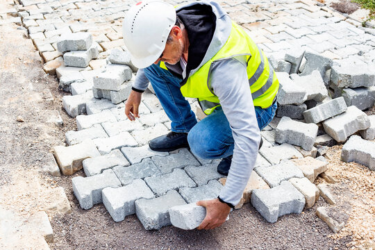 construction worker in white safety helmet and yellow reflective vest placing paving stones on a ground for new pavement road outdoors, construction and industry concept 