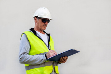 Man engineer or architect in black sunglasses in white safety helmet and yellow reflective vest is writing on clipboard against beige wall, industry and construction concept 