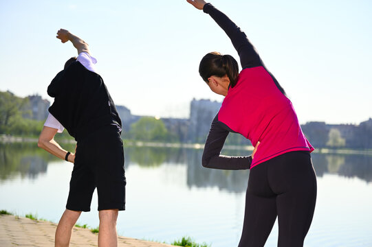 The stunning photo captures a young couple as they warm up, stretch and train near the serene lake. Fit and active young couple dedicated of a dynamic stretching routine by the tranquil urban lake.