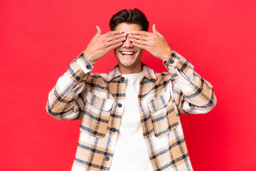 Young caucasian handsome man isolated on red background covering eyes by hands and smiling