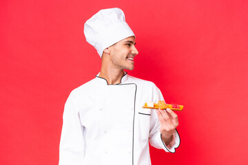 Young caucasian chef man holding sashimi isolated on red background looking side