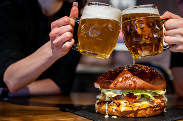 man and woman hands clinking with glasses of light beer at the pub or bar and very big burger