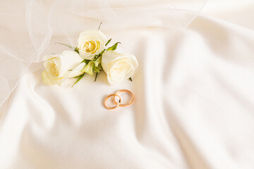 Two gold wedding rings on a beige satin background with live tea roses and part of the bride's...