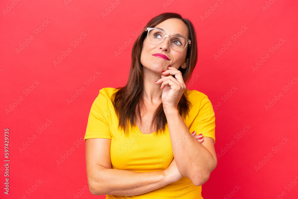 Wall mural Middle-aged caucasian woman isolated on red background having doubts while looking up