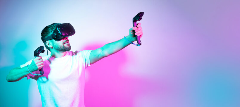 Concept modern hobby. Bearded millennial man in VR glasses plays in simulation figth and using controllers in gradient neon background. interactive online game in a virtual world. Copy space, banner