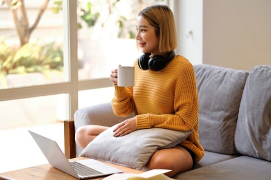 young asian woman student with headphones using laptop in a video call or online class while sitting on sofa at home