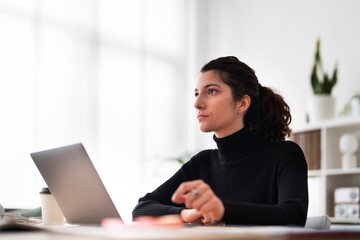 Positive young female sitting at table with laptop and looking at computer screen while working in modern office
