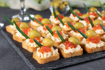 Festive canapes with red caviar and olives on croutons of white bread on slate board on gray background