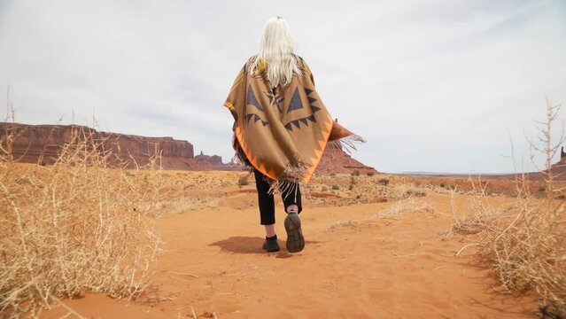Young woman tourist wrapped in brown orange blanket with traditional pattern with blond hair, steps along path at Monument Valley landscape. Cute girl in beautiful cinematic Monument Valley park