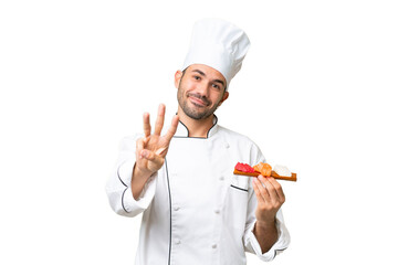 Young caucasian chef holding a sushi over isolated background happy and counting three with fingers