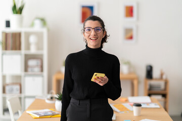 Positive young female in casual clothes smiling and looking at camera while sitting at table working on project at computer in bright modern location