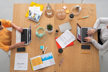 Top view of crop anonymous female coworkers in casual clothes sitting at table with papers and working on project on laptop