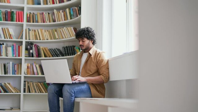 Young student using laptop computer to type for online e-learning in campus library space. A handsome male is preparing for the examination period at the university in the classroom sitting on a bench
