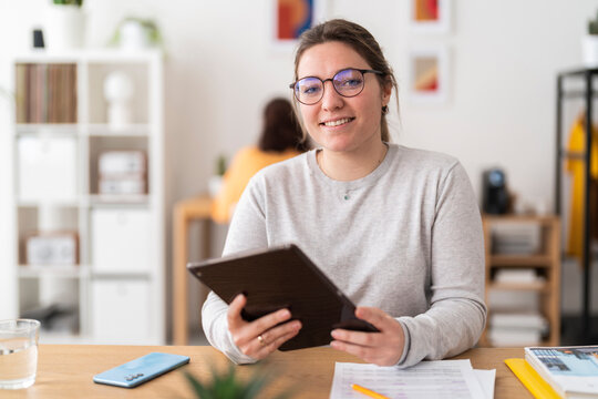 Positive woman in casual clothes smiling and looking at camera while sitting at table with tablet and working in modern workspace