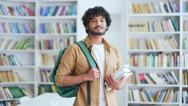 Portrait of young smiling university student holding books, textbooks, looking at camera while standing in campus library space. Happy male with backpack in college classroom preparing for exam period