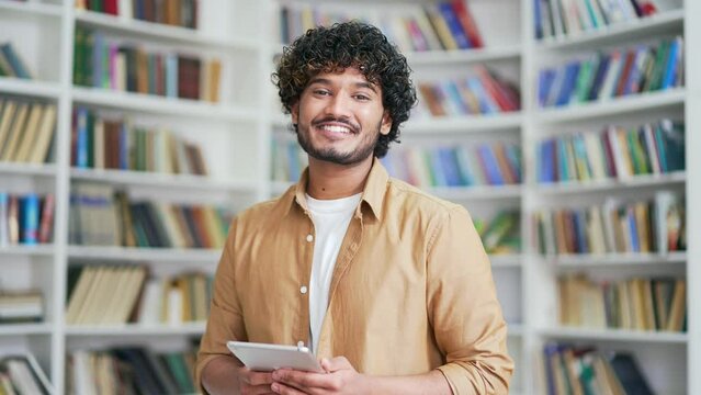 Portrait of young smiling university student holding tablet and looking at camera while standing in campus library space. Happy positive friendly male with backpack in college classroom posing