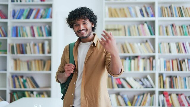Portrait of a happy young university student smiling, looking at the camera and calling to him with a hand gesture. College teacher calling for learning and knowledge standing in campus library space
