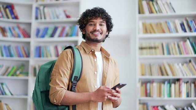 Portrait of young smiling university student holding smartphone and looking at camera while standing in campus library space. Happy positive friendly male with backpack in college classroom posing