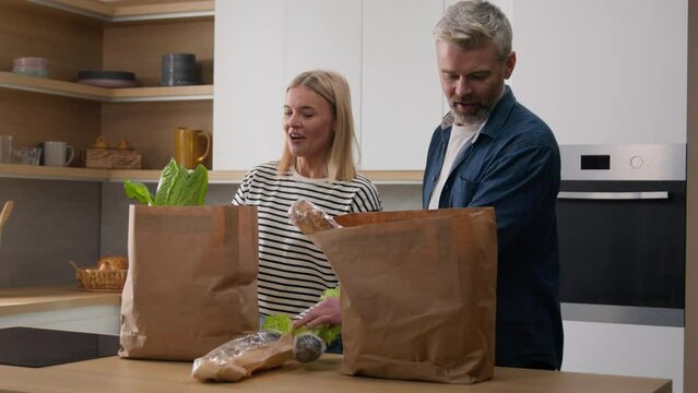 Caucasian middle-aged couple happy family adult wife and husband woman and man at home kitchen unpack shopping bag packages products with lettuce cabbage apple juice water bread food delivery talking