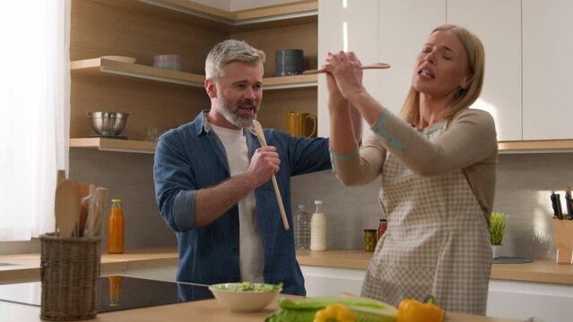 Middle-aged couple happy family cheerful wife and husband singing song having fun fooling around at kitchen together cook fresh vegetables salad adult woman and man sing in kitchenware utensil spoons