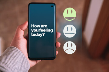 Woman's hand holds a smartphone while choosing a smile happy face on screen about how she feels. Feedback and rating positive. Mental health assessment. Wellness and think positive.
