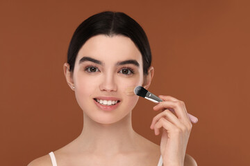 Teenage girl applying foundation on face with brush against brown background