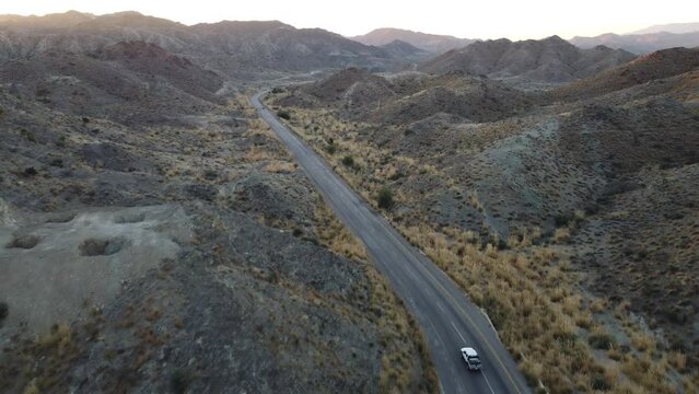 Aerial drone following shot over a white car moving along a winding RCD road through mountainous terrain in Balochistan during evening time.