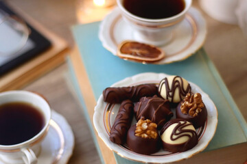 Books, reading glasses, e-reader, plate of chocolate pralines, bowl of cookies, cups of tea and lit candles on the table. Selective focus.
