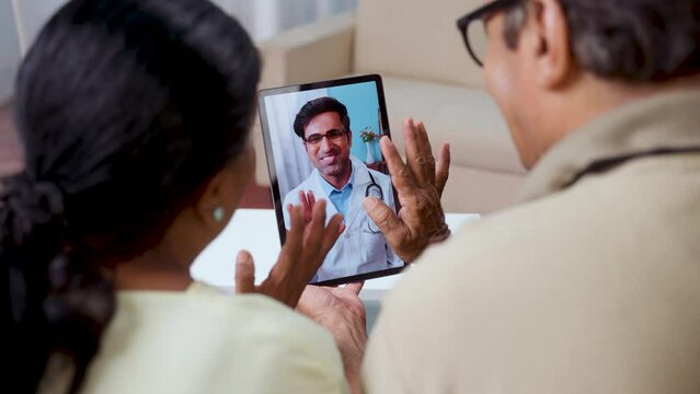 Shoulder shot of Indian Senior couple taking advice from doctor during online video call from tab at home - concept of remote health care, virtual consultation and ehealth