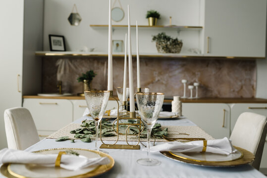 Festive table setting with plate, golden cutlery set and eucalyptus leaves, candles. Dinner table decorated for cozy fall holiday gathering in kitchen. Modern panel kitchen interior with countertop. - Powered by Adobe